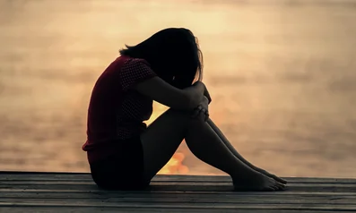 Female on a dock by water with her head down and her arms crossed resting on her knees, overcome by feelings of trauma.